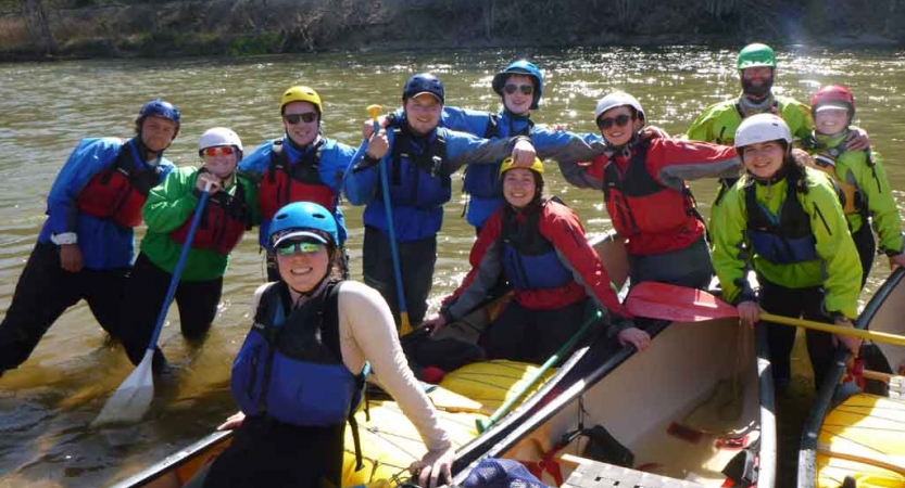 A group of people wearing safety gear stand in or near knee-deep water, and beside two canoes, and pose for the photo